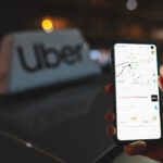 Uber app displayed on smartphone held in hand in front of Uber taxi sign on top of a car at night with city lights background with soft focus. Taking a cab concept. Warsaw, Poland - October 23, 2021